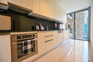 A modern kitchen with stainless steel appliances, a black countertop, and a view of a high-rise building through large windows. - Luxury Marina 1-Bedroom - 5-star Hotel Amenities (Dubai)