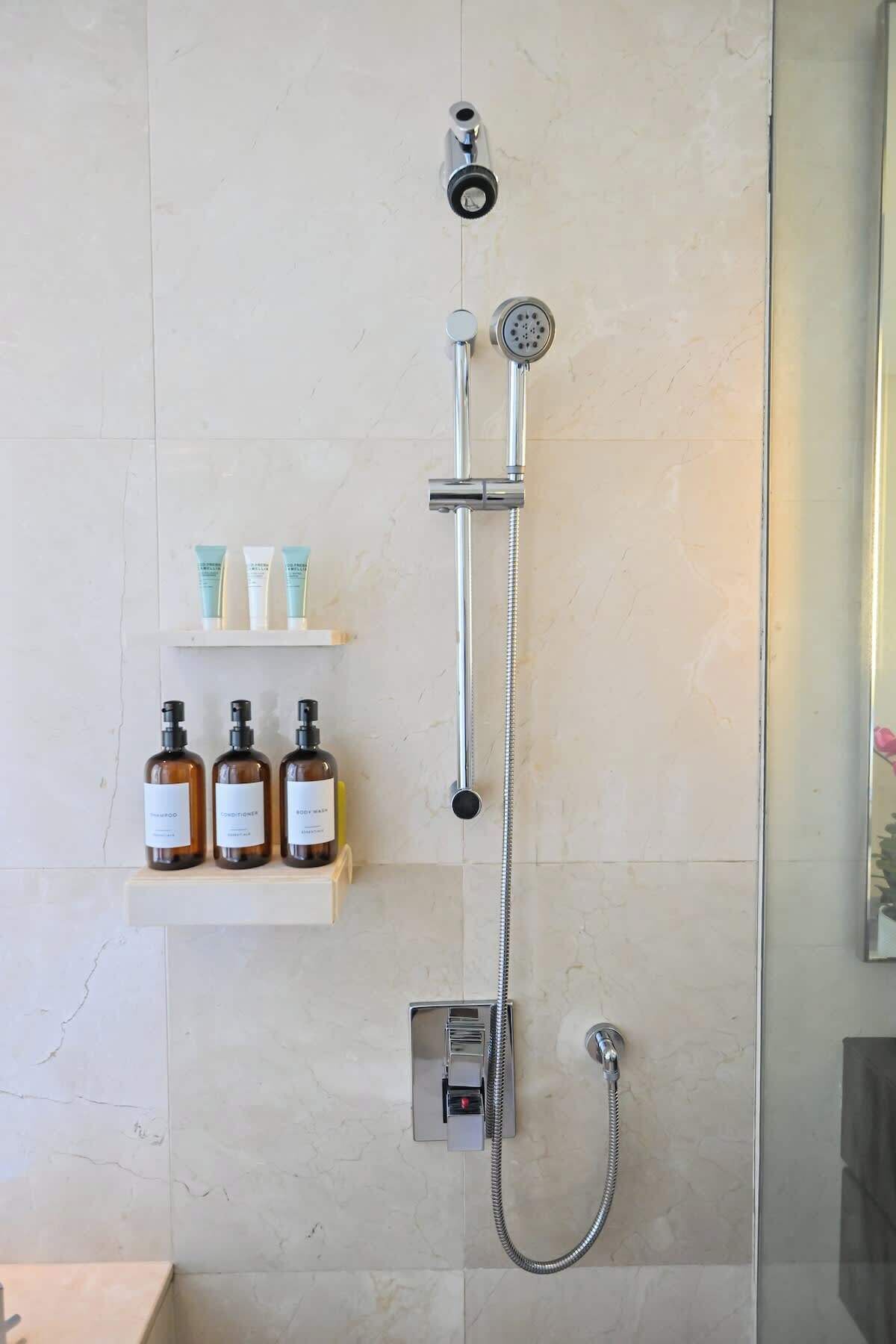 A bathroom with a wall-mounted shower, a handheld showerhead, and a handheld shower attachment. There are three bottles of shampoo and conditioner on a shelf below the shower.