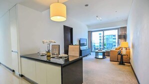 A modern hotel room with a kitchen area, a dining table, a sofa, and a large window offering a city view. - Luxury Marina 1-Bedroom - 5-star Hotel Amenities (Dubai)