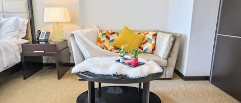 Une chambre d’hôtel avec un lit, une table de chevet avec une lampe, un canapé avec des coussins à motifs et une table ronde recouverte d’une housse blanche et moelleuse.