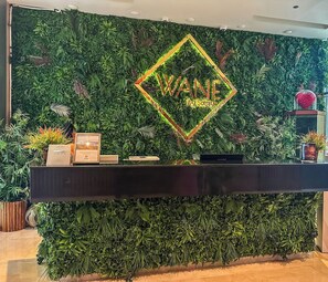 Balcão de recepção com parede de fundo verde, um letreiro neon que diz 'WANE' e uma pequena planta em vaso à direita.