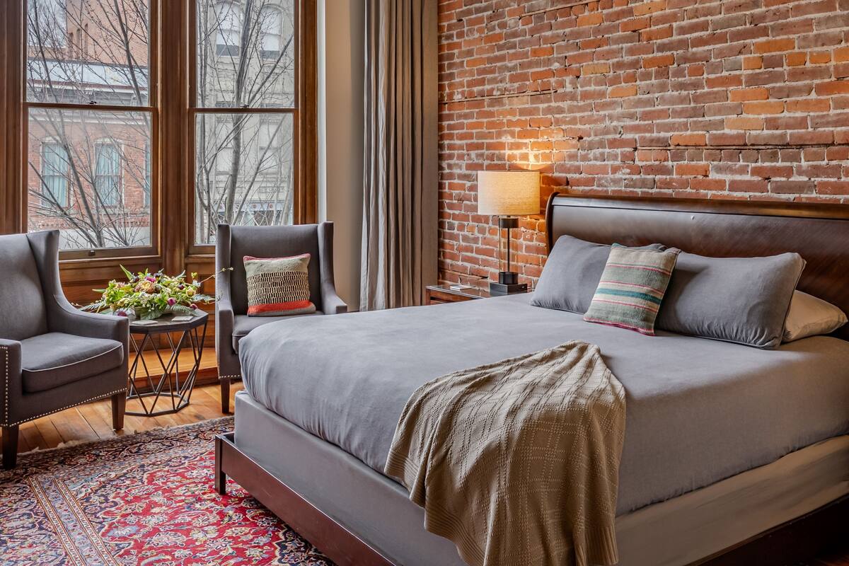 A bedroom with a large bed, two armchairs, a small side table with a lamp, and a view of the city through large windows.