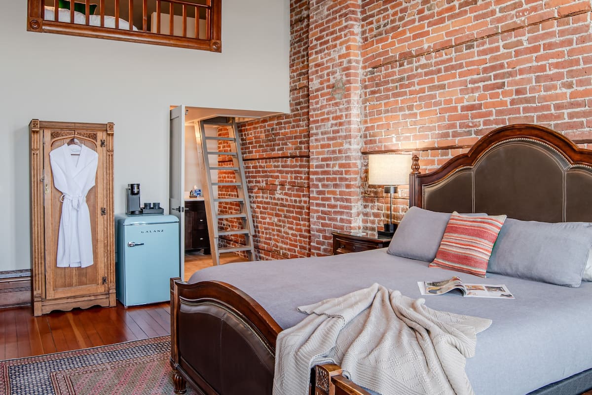 A bedroom with a brick wall, a wooden bed, a wardrobe, and a small blue appliance.