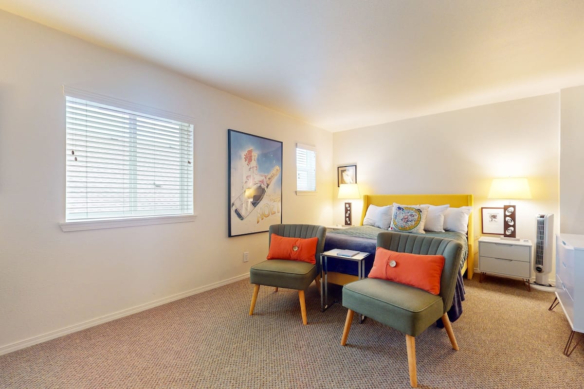 A bedroom with a bed, two chairs, a nightstand, a lamp, and a window with blinds.