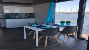 Un comedor moderno con una mesa blanca y sillas negras, una cocina con gabinetes blancos y vistas al agua con barcos.