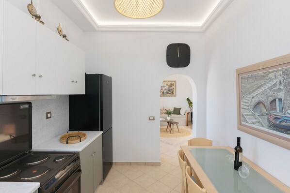A modern kitchen with a stove, refrigerator, and white cabinets. There is a dining area with a table and chairs, and a painting on the wall. - Ocean Caldera View Villa by Dalmyra Estate (Santorini)