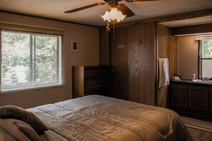 Un dormitorio con cama, ventilador de techo, ventana con persianas, puerta de madera y baño con lavabo y espejo.