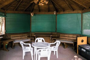 Un espacio de estar al aire libre cubierto con bancos de madera y sillas de plástico blanco, una mesa y un armario de madera.