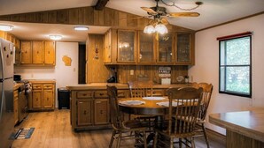 Una cocina con armarios de madera, una mesa de comedor con sillas, un ventilador de techo y una ventana con vista a árboles.