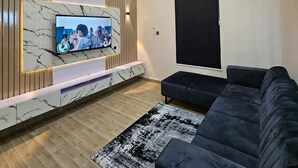 A modern living room with a flat-screen TV mounted on a marble wall, a black sectional sofa, and a patterned rug. - Luxury 4 Bed Duplex + PS5/Snooker/Tennis Lekki (Lekki)