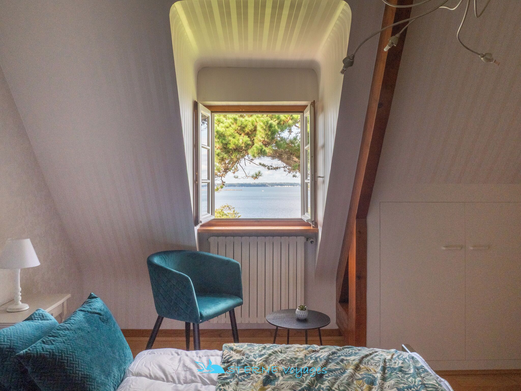 Un dormitorio con ventana que da a una masa de agua, una silla color turquesa, una mesita y una cama con colcha estampada.