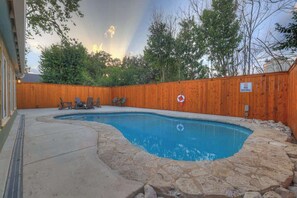 A backyard with a swimming pool, stone patio, wooden fence, and outdoor furniture.