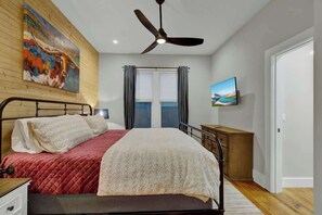 Ein Schlafzimmer mit einem Bett, einem Deckenventilator, einem Gemälde, einem Fernseher und einem Fenster mit Vorhängen.