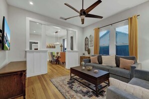Ein Wohnzimmer mit einer Couch, einem Couchtisch und einem Deckenventilator. Im Hintergrund ist eine Essecke mit einem Kronleuchter zu sehen.