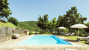 Une piscine rectangulaire bordée de pierres, entourée de chaises longues et de parasols, avec en arrière-plan un paysage de arbres et de collines.