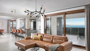A modern living room with a leather sectional sofa, a round coffee table, and a chandelier. There is a dining area with bar stools and a kitchen in the background. The room has large glass doors leading to a balcony with a view of the sea. - Infinity Blue and Golden Sky (Vir)