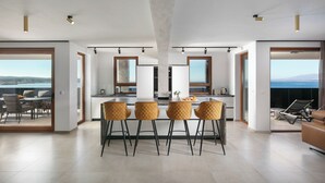 A modern kitchen with a central island, bar stools, and large windows offering a view of the sea. - Infinity Blue and Golden Sky (Vir)