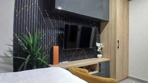 A modern bedroom with a bed, a TV mounted on a wall, a small shelf with a vase, and a plant. - Grey Courts (Accra)