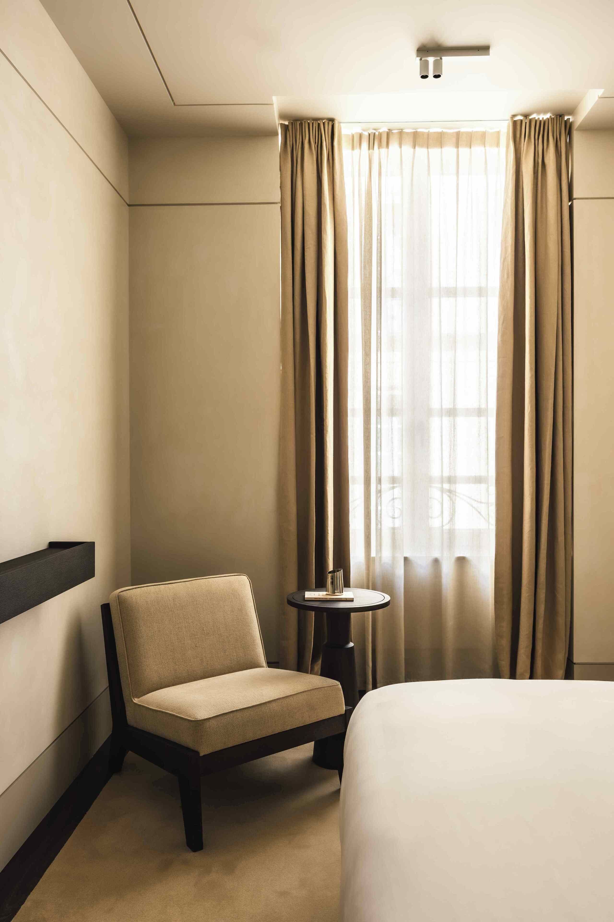 A hotel room with a bed, a chair, a small table, and a window with curtains.
