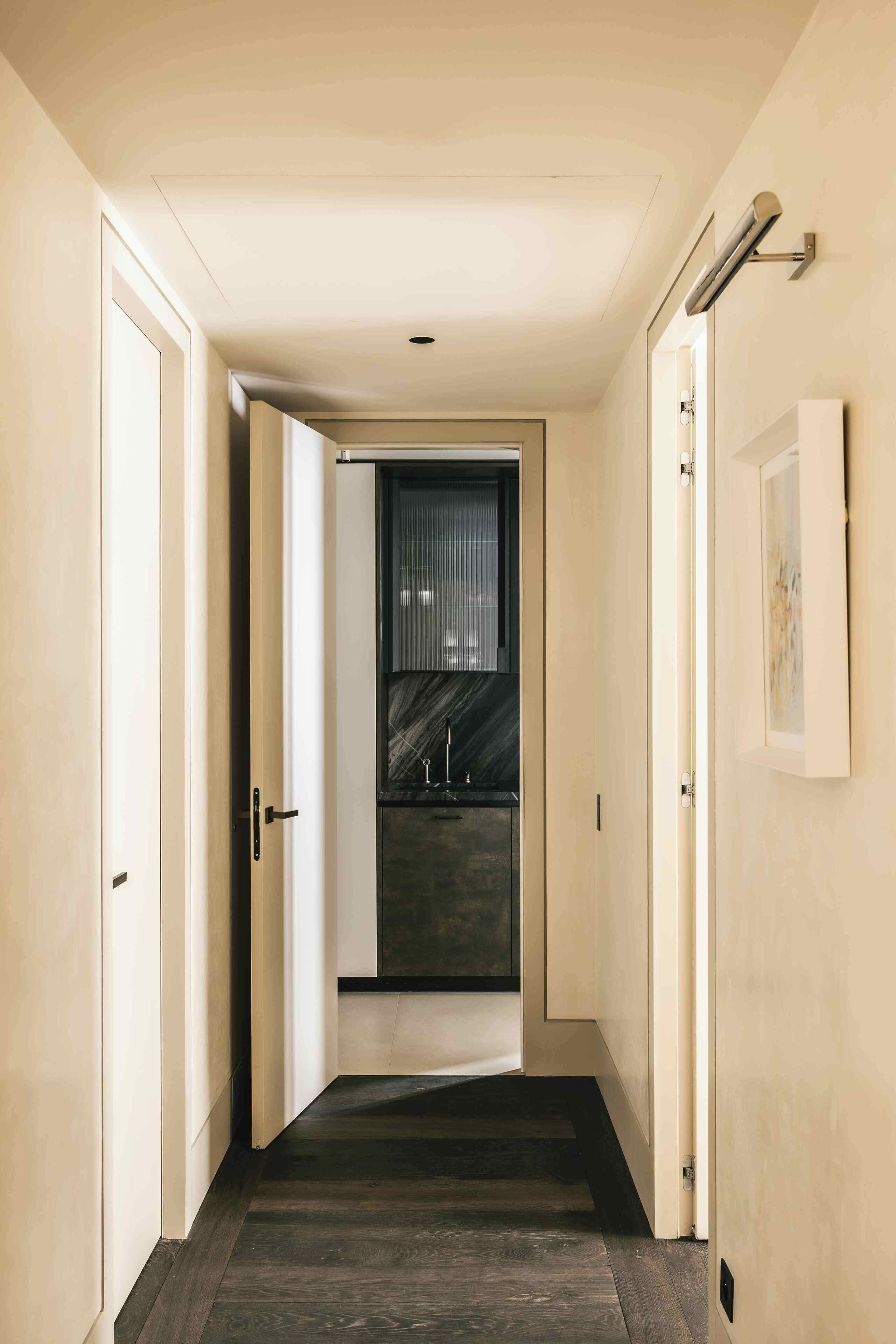 Un couloir doté d’un plancher en bois foncé, de murs de couleur beige et d’une porte donnant sur un espace cuisine avec des plans de travail sombres et une fenêtre.
