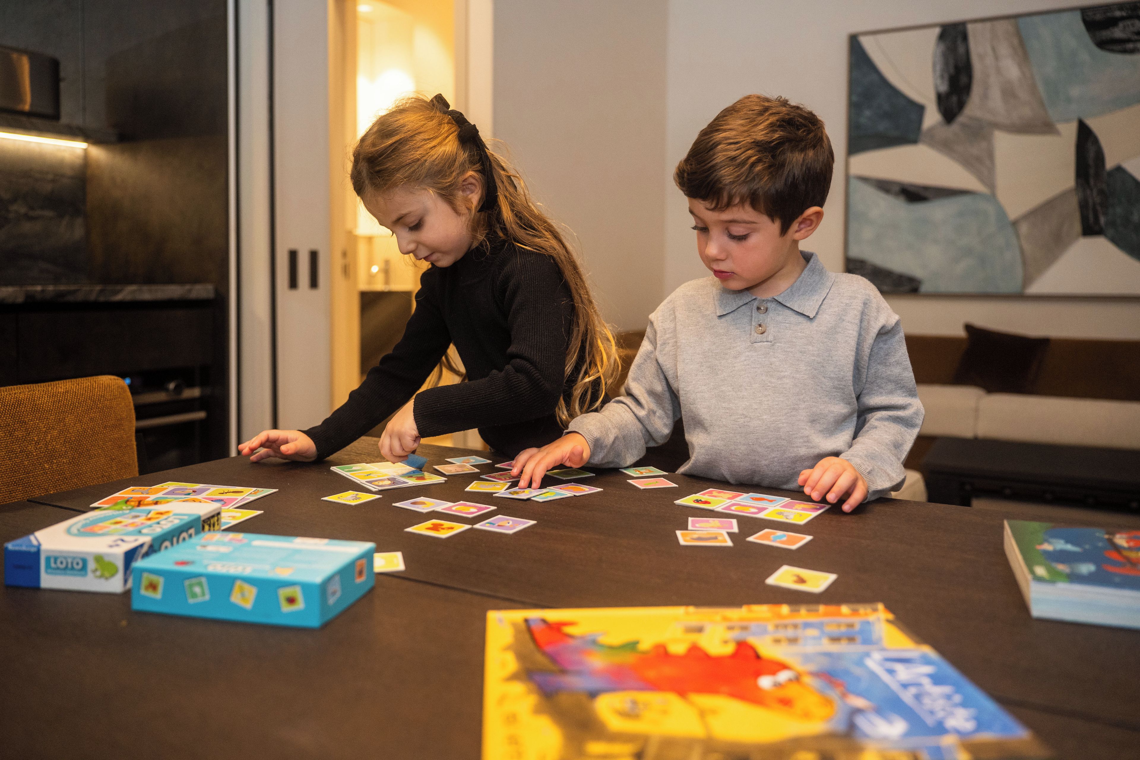 Zwei Kinder spielen Kartenspiele an einem Tisch, im Hintergrund steht eine Couch und ein Gemälde.