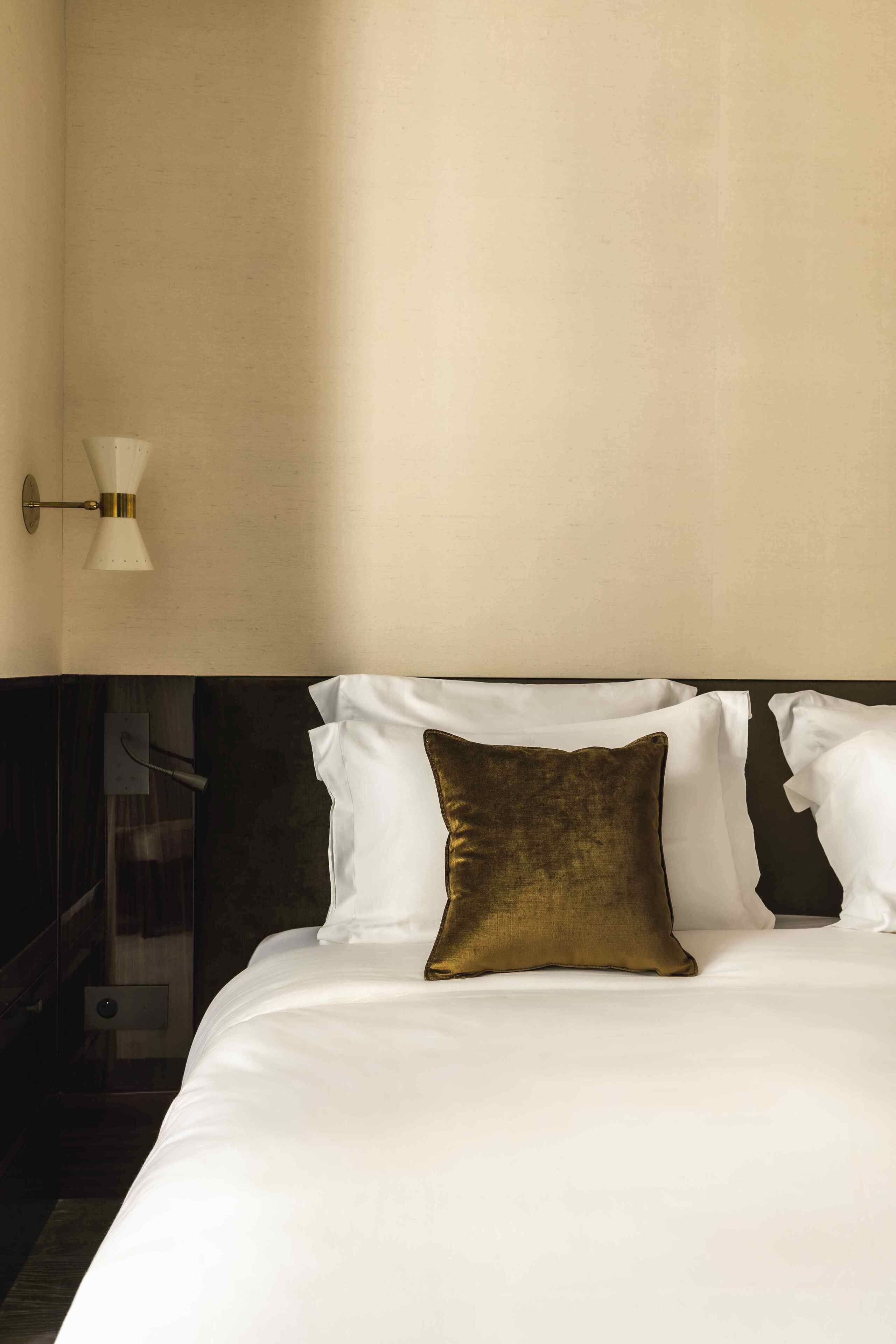 Een netjes opgemaakt bed met witte en gouden kussens, een wandlamp en een donker hoofdbord.