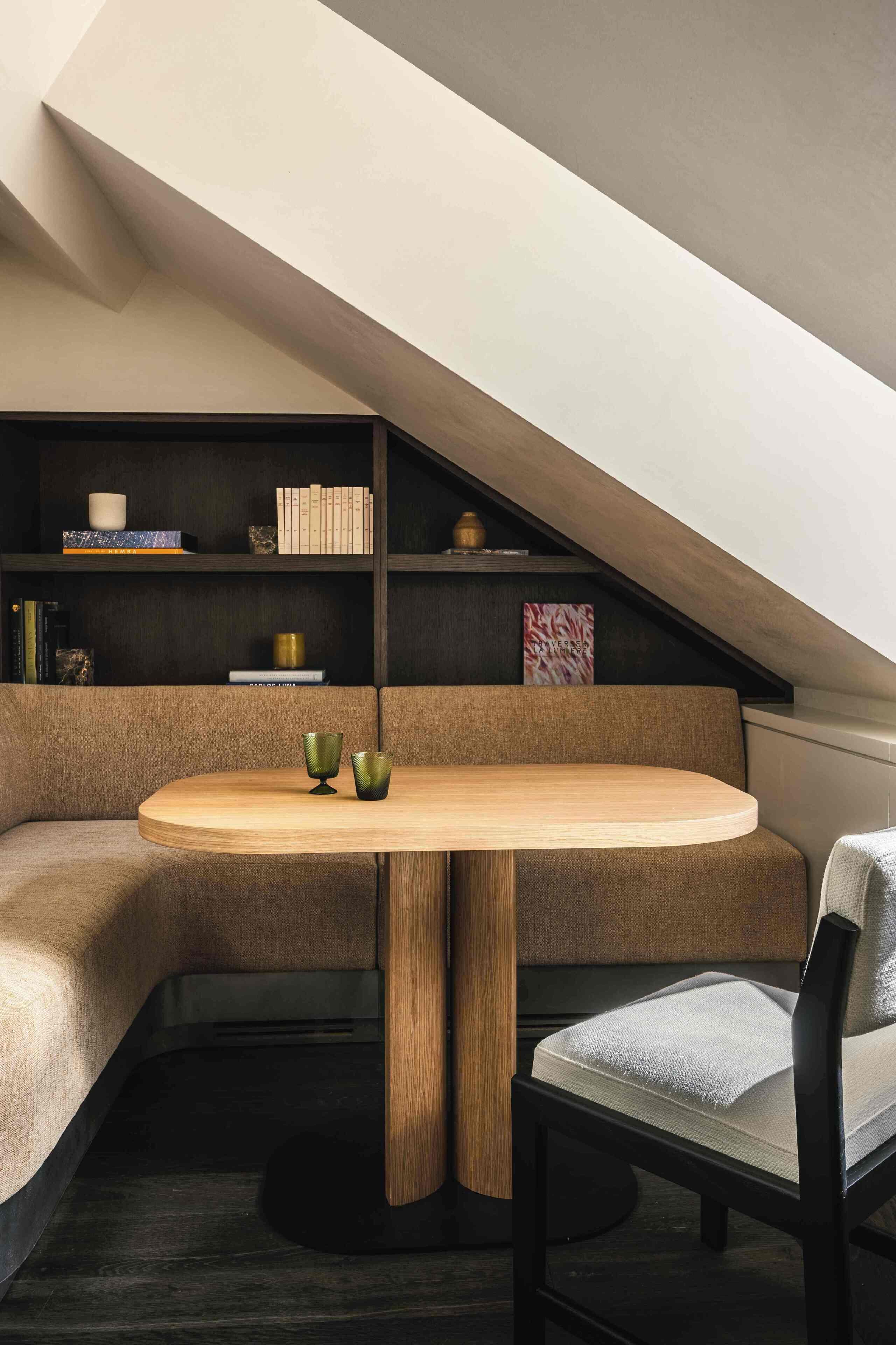Un coin accueillant avec une table ronde en bois, un banc rembourré et une bibliothèque remplie de livres et d’objets décoratifs.