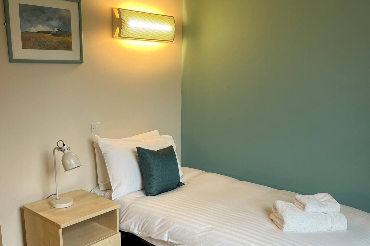 A neatly made bed with white linens and two pillows, a bedside lamp, a small wooden nightstand with a lamp, and a framed picture on the wall.