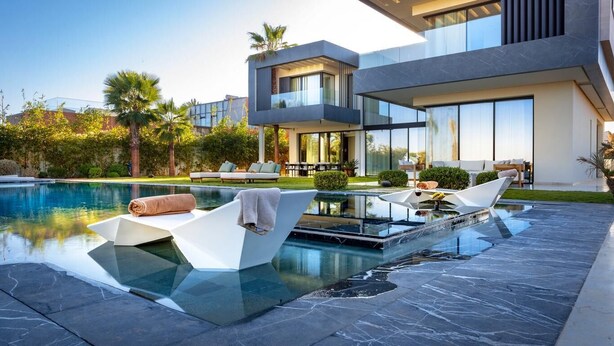 Un área de piscina moderna con un jacuzzi de vidrio, sillones de descanso y un jardín cuidado.