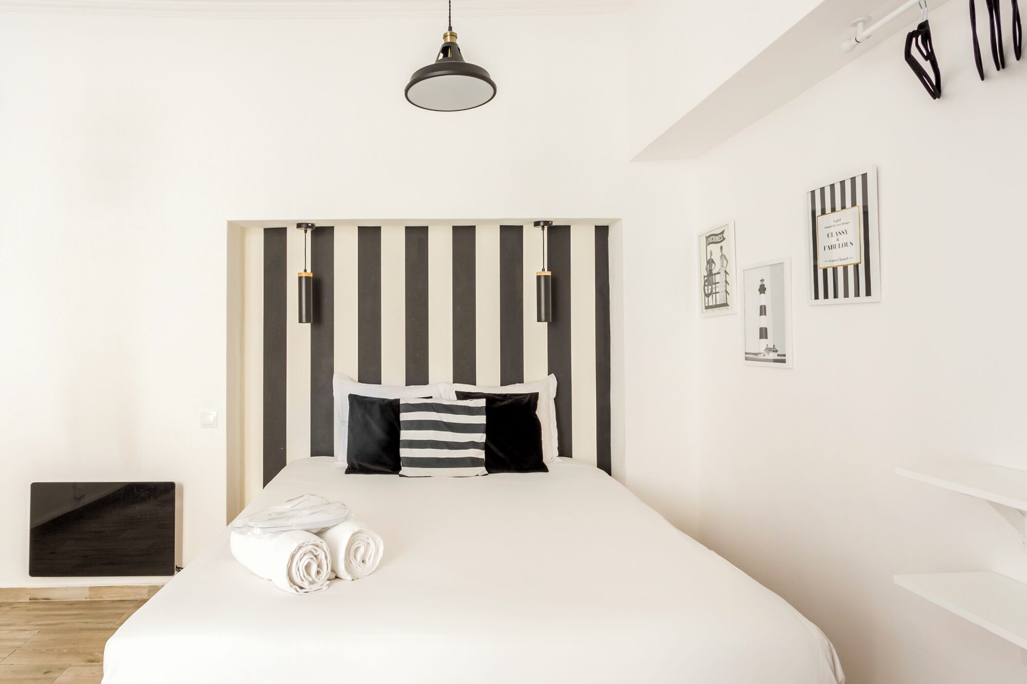 A modern hotel room with a large bed, a headboard with vertical stripes, a hanging pendant light, and a small bedside table with a lamp.