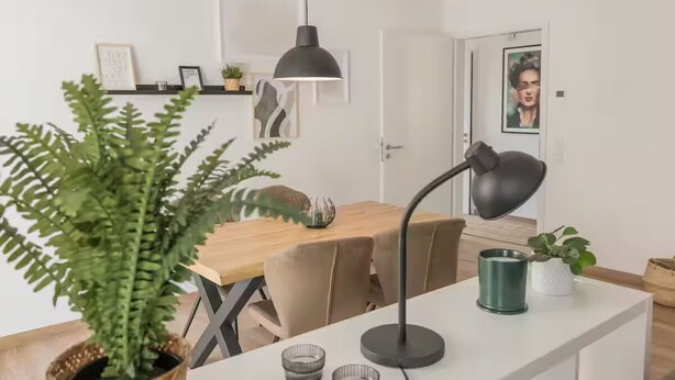 A modern dining area with a wooden table, chairs, a black pendant light, and a potted fern.