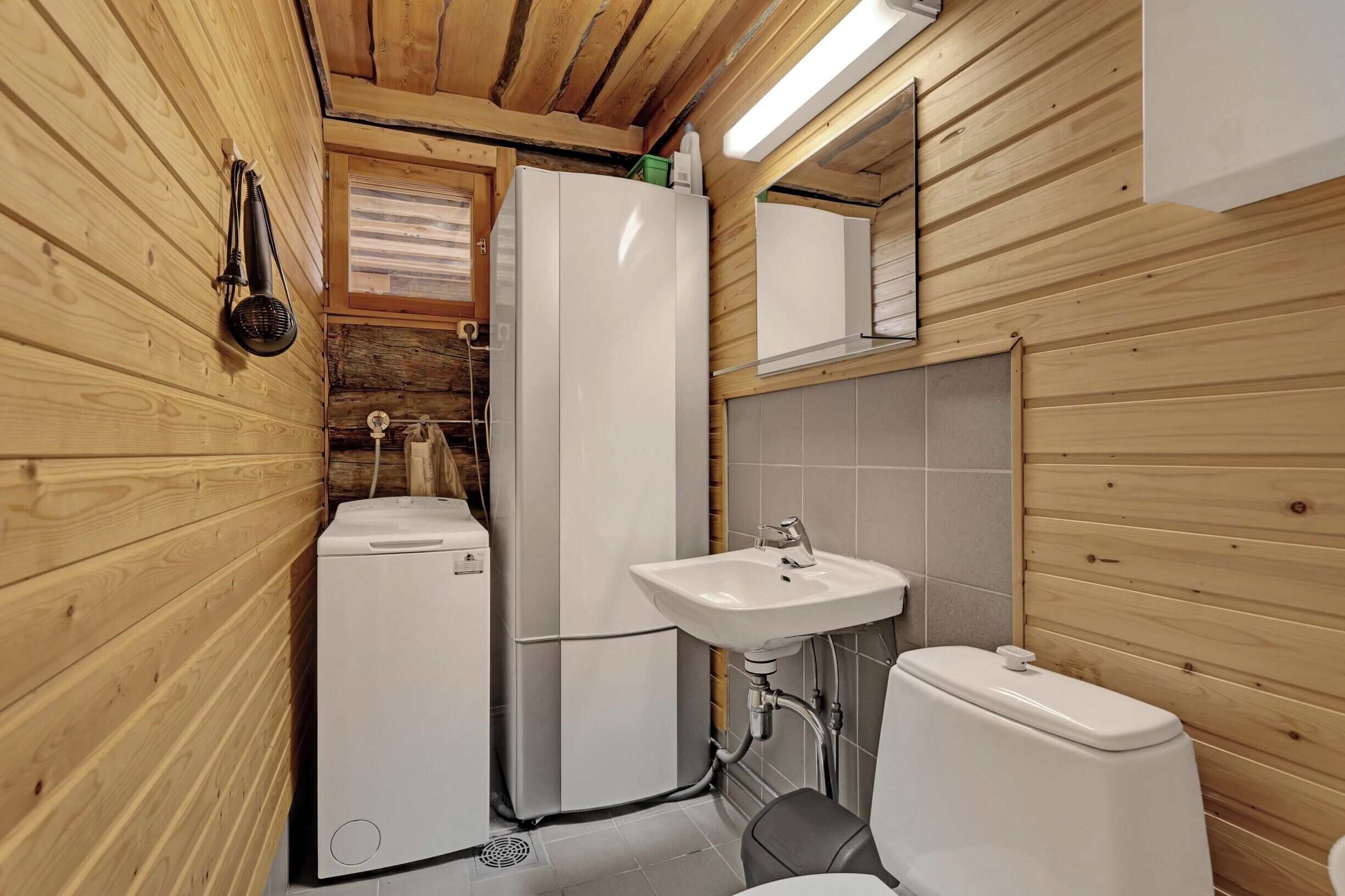 A compact bathroom with wooden walls, a white sink, a toilet, and a small refrigerator.