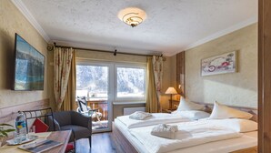 A hotel room with a wooden bed, a desk with a chair, a table with a bottle and glasses, a window with curtains, and a painting on the wall. - The Rotspitz / Achensee (Tirol)