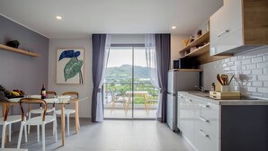 A modern kitchen with white cabinets, a dining area with a table and chairs, and a view of greenery through large windows. - Thai me up the Tower (Chalong)