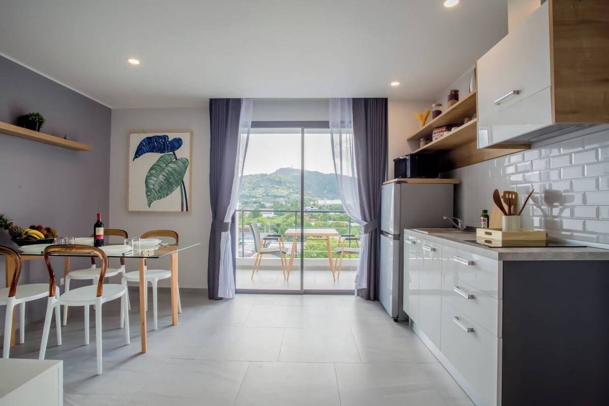 A modern kitchen with white cabinets, a dining area with a table and chairs, and a view of greenery through large windows.