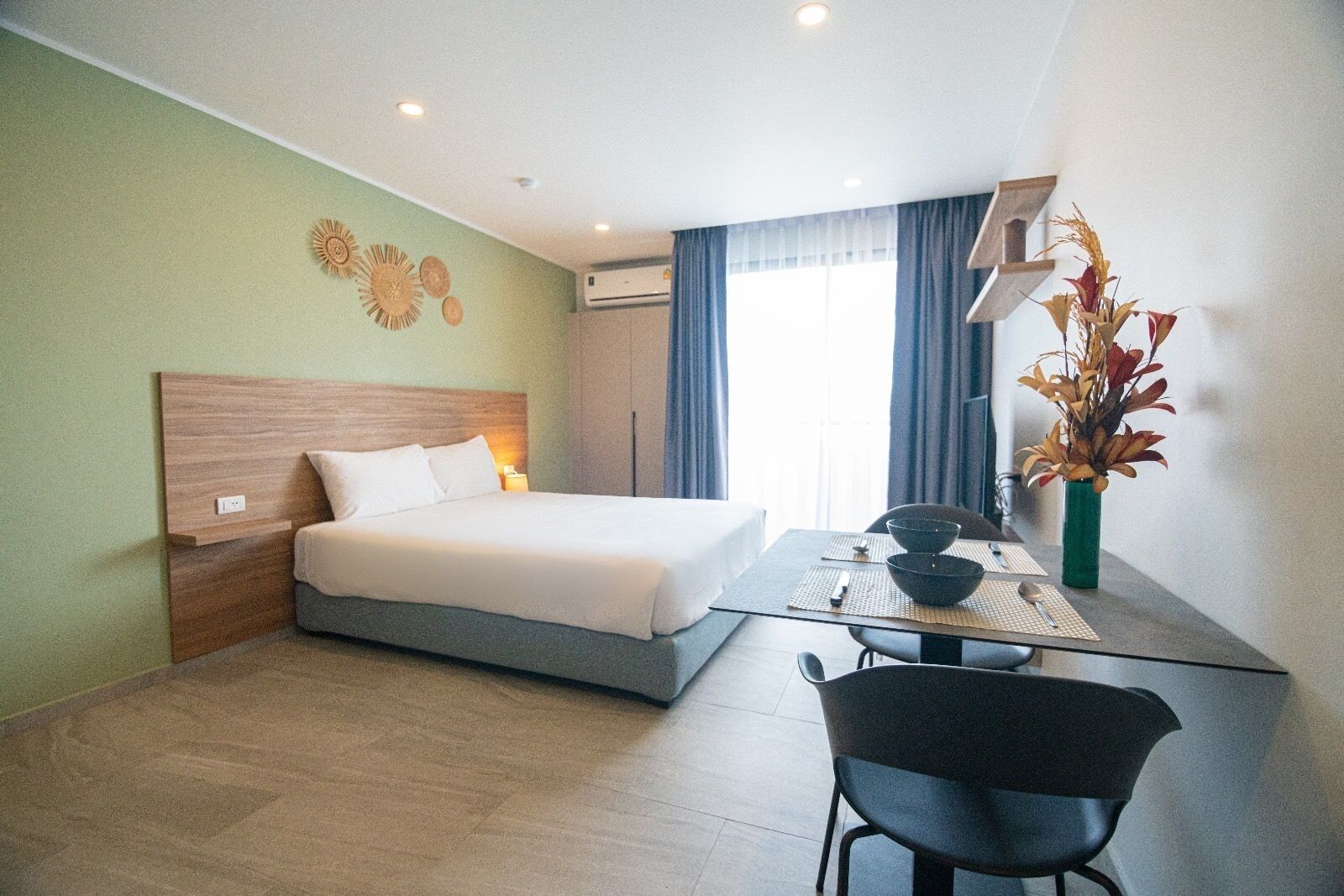 Ein Hotelzimmer mit einem Bett, einem Schreibtisch mit Stuhl, einer Vase mit Blumen und einem Fenster mit Vorhängen.