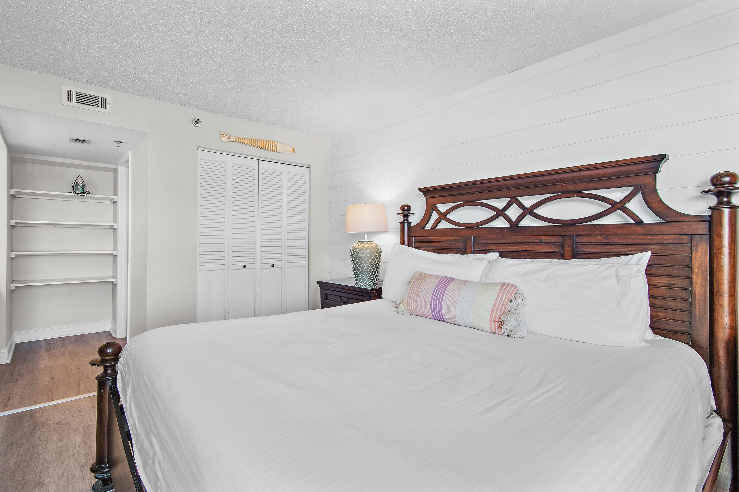 A bedroom with a wooden headboard, a bed with white bedding, a nightstand with a lamp, and a white wardrobe with louvered doors.