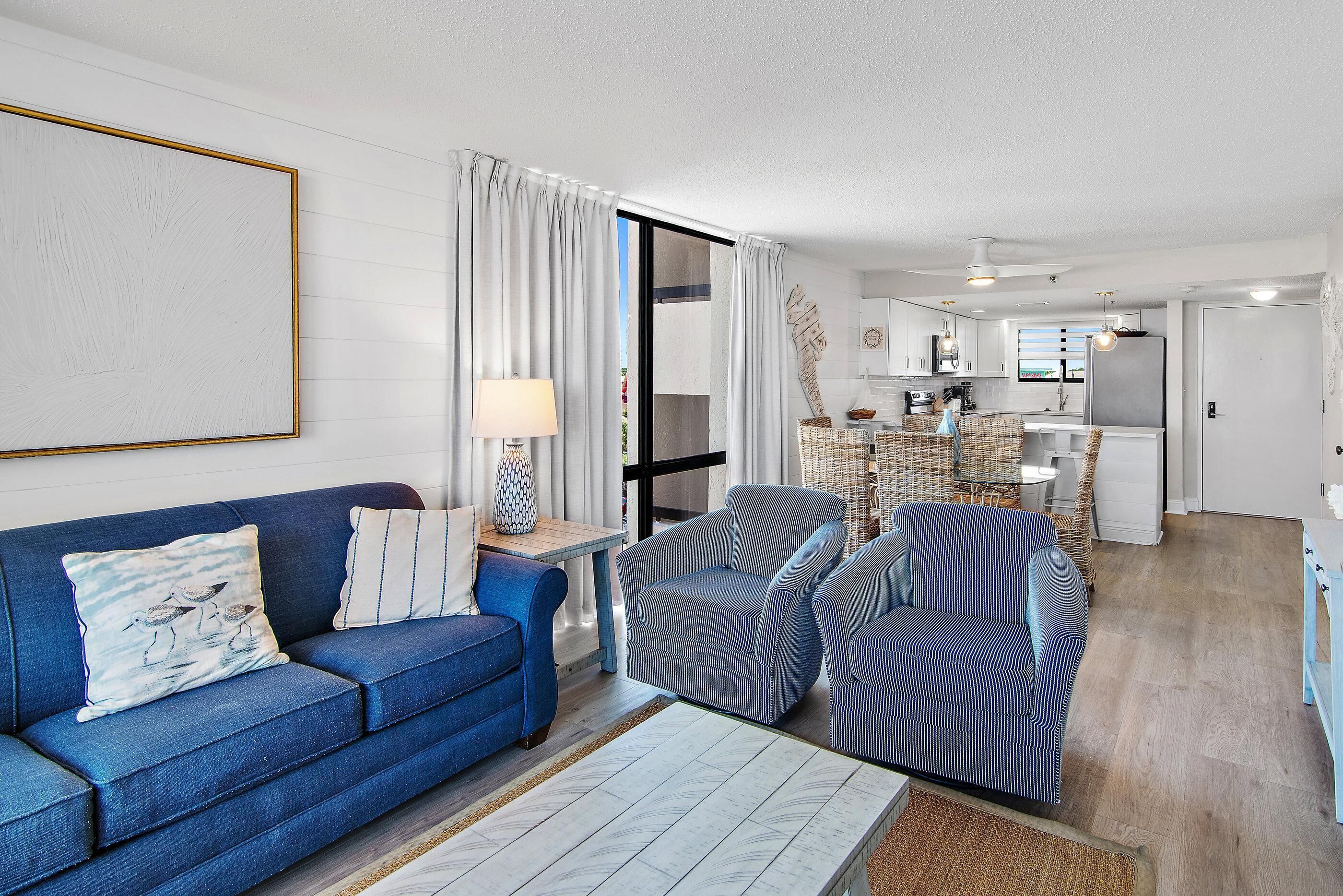 A modern living room with a blue sofa, striped armchairs, a coffee table, and a dining area with wicker furniture.