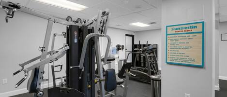 A gym with various exercise machines, a wall-mounted sign with rules, and a door leading to another room.