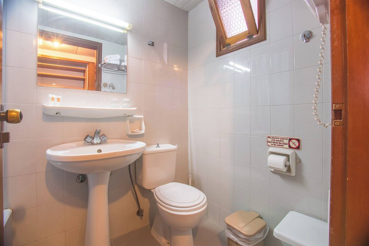 Un baño con lavamanos, inodoro y bidé blancos. Hay un espejo sobre el lavamanos y un letrero en la pared.