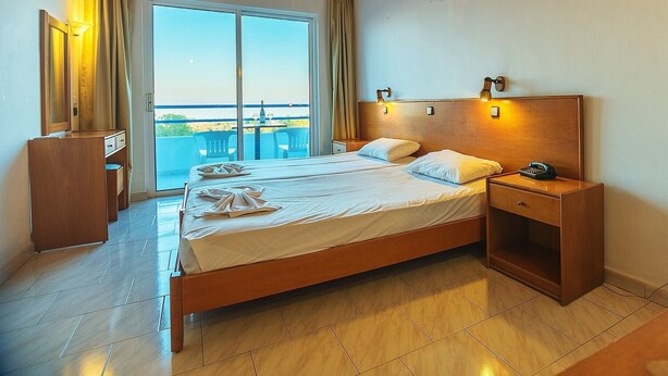 A hotel room with a wooden ceiling, a large bed with white linens, a nightstand with a phone, and a view of the beach through the window. - Celadi vista (Rhodes)