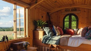 A cozy cabin interior with a bed, a window overlooking a forest, a small table with a plant, and a guitar.