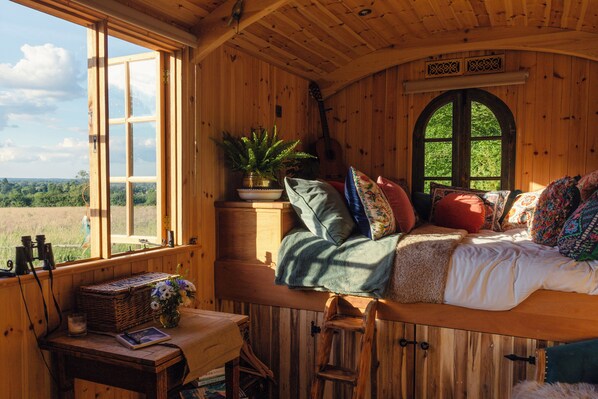 A cozy cabin interior with a bed, a window overlooking a forest, a small table with a plant, and a guitar.