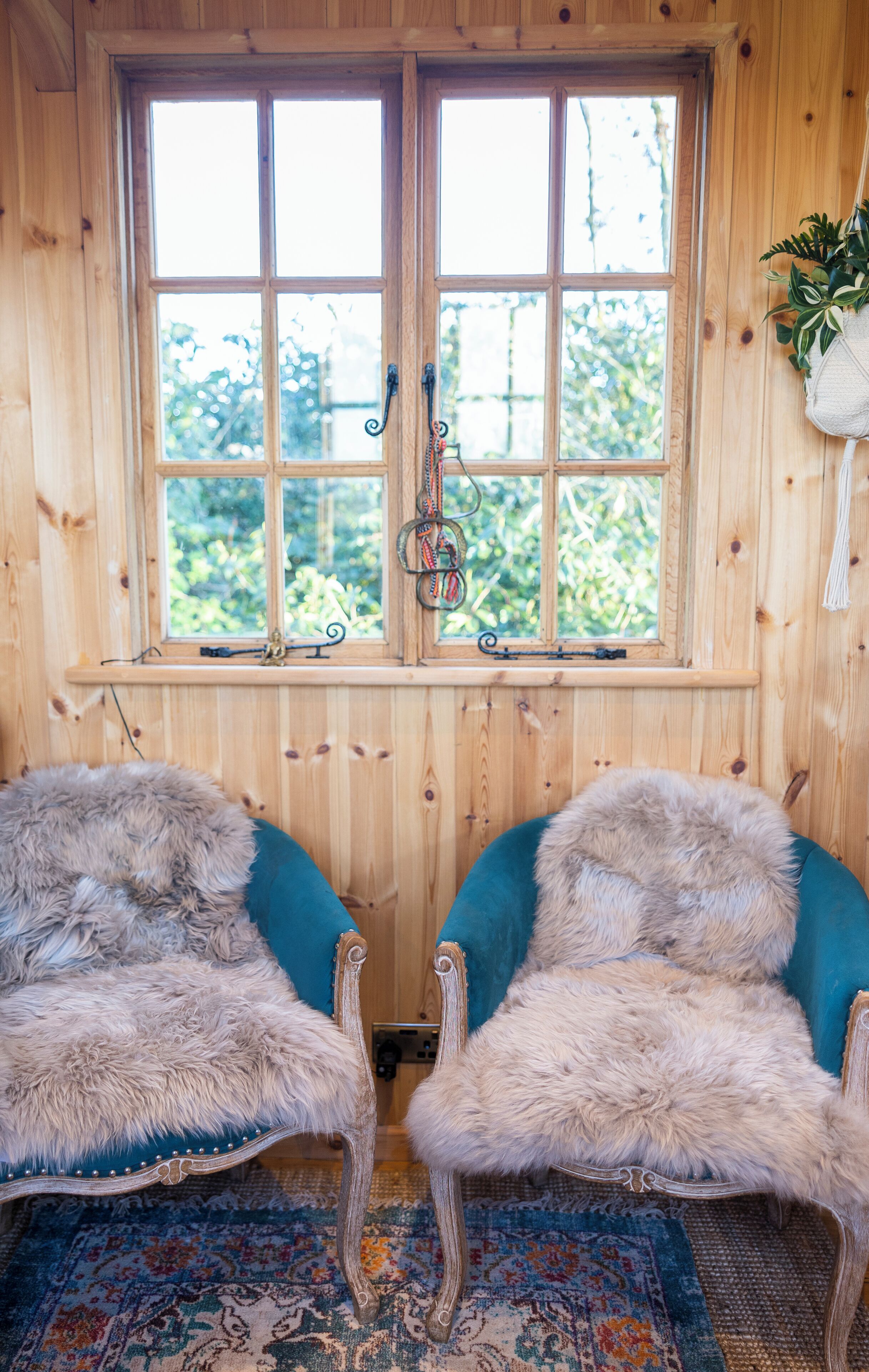 A room with wooden walls, a large window with a view of greenery, two chairs with fur upholstery, and a patterned rug.