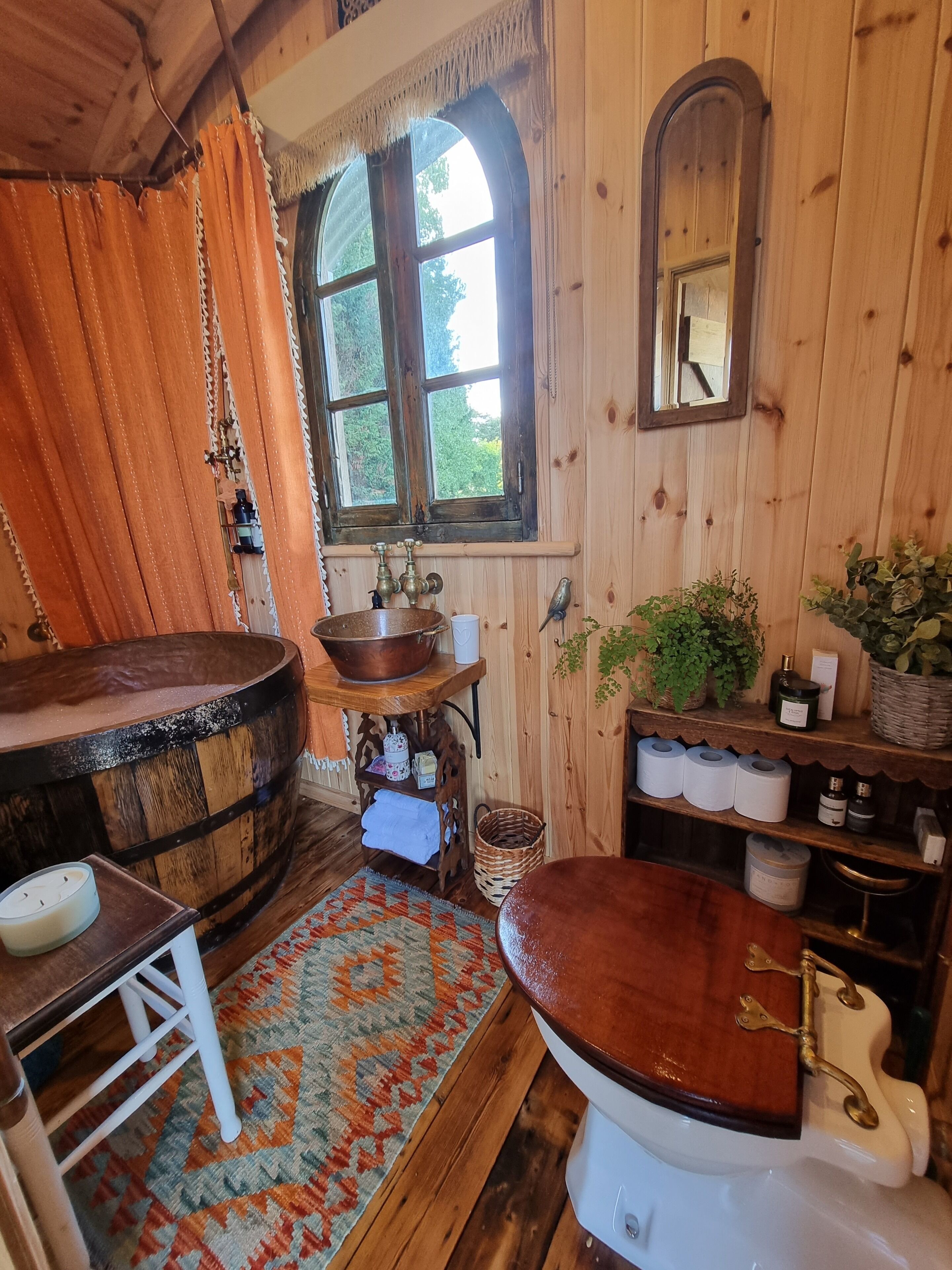 A rustic bathroom with a wooden bathtub, a white toilet with brass fixtures, a wooden vanity with a mirror, and a window with curtains.