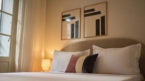 En pænt redt seng med hvide og brune puder, en sengetårnslampe og to abstrakte malerier på væggen.