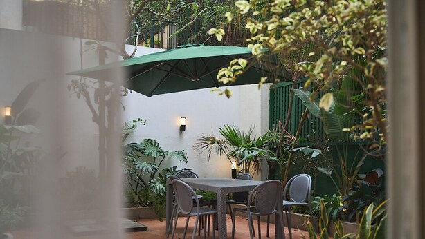 En udendørs terrasse med et bord og stole, omgivet af forskellige planter og en grøn parasol.