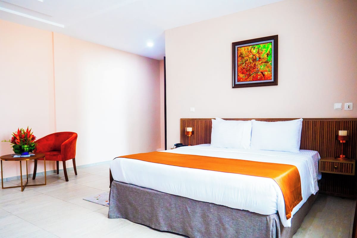 A hotel room with a large bed, a red chair, a small table with a vase, and a painting on the wall.