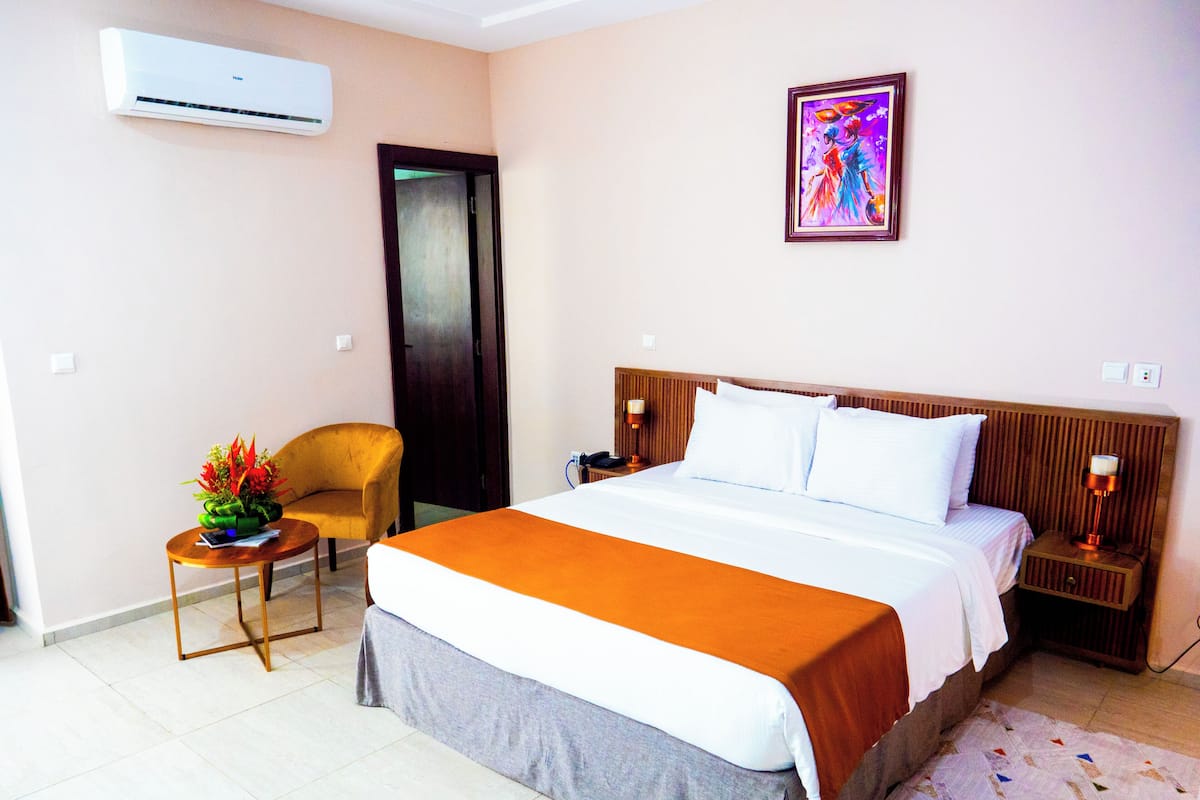 A hotel room with a bed, a chair, a nightstand, a lamp, an air conditioner, and a painting on the wall.