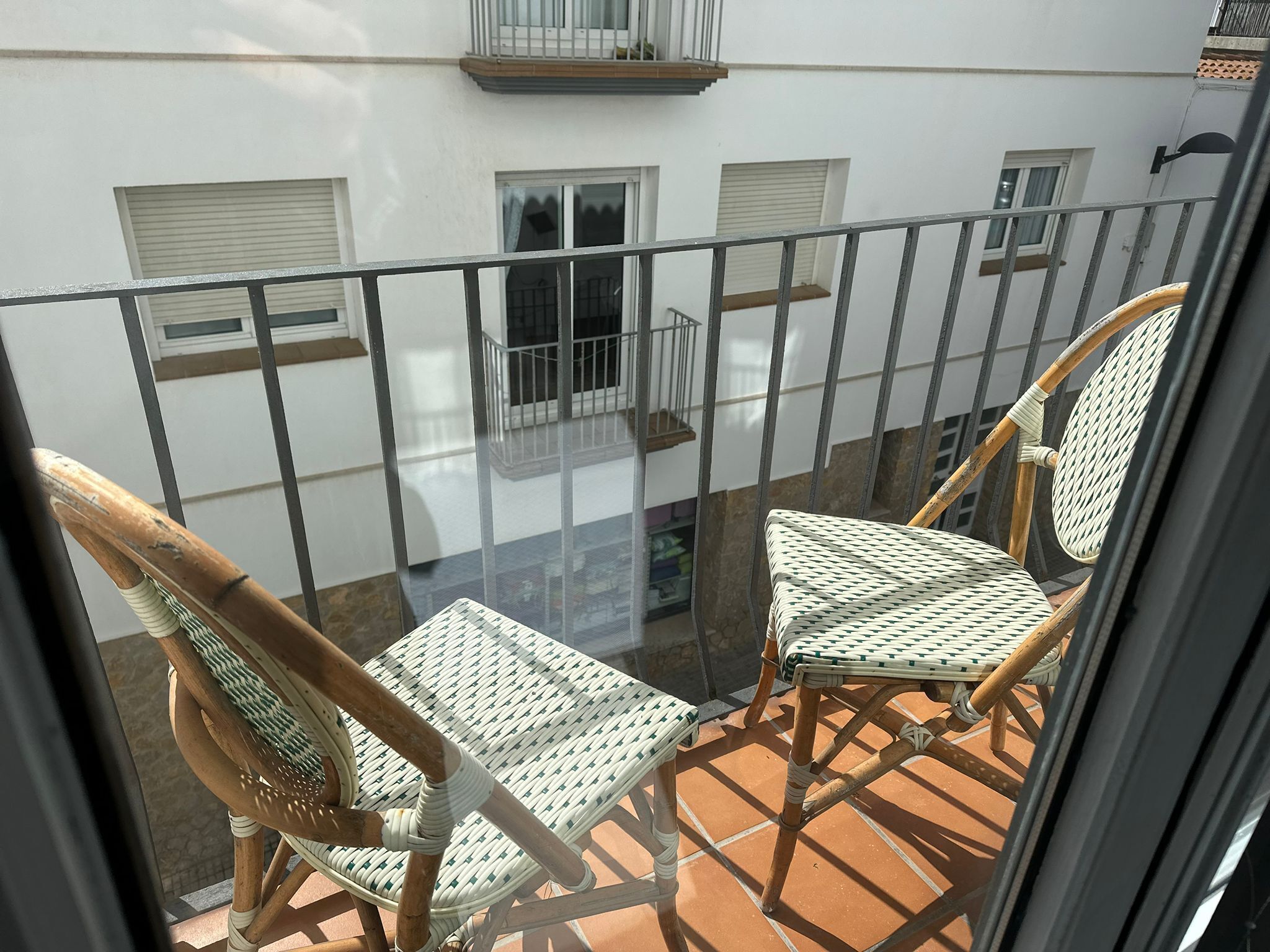 A balcony with two wicker chairs, a glass table, and a view of a building with windows and a balcony railing.